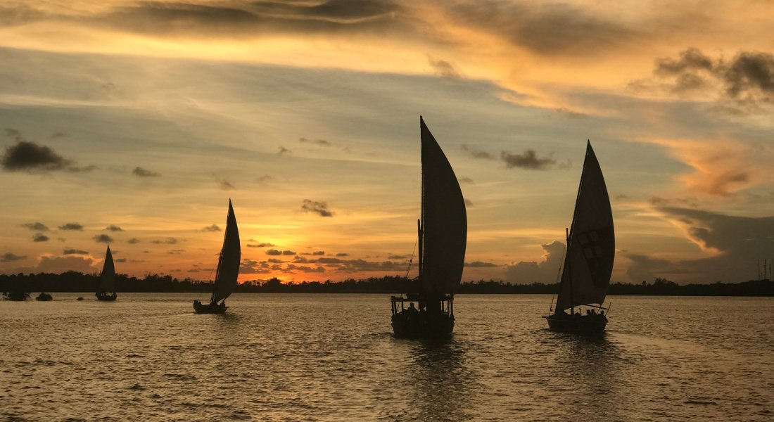 Elegant dhow sailboats in Lamu, Kenya, glide across the ocean at sunset, painting the horizon with warm hues.
